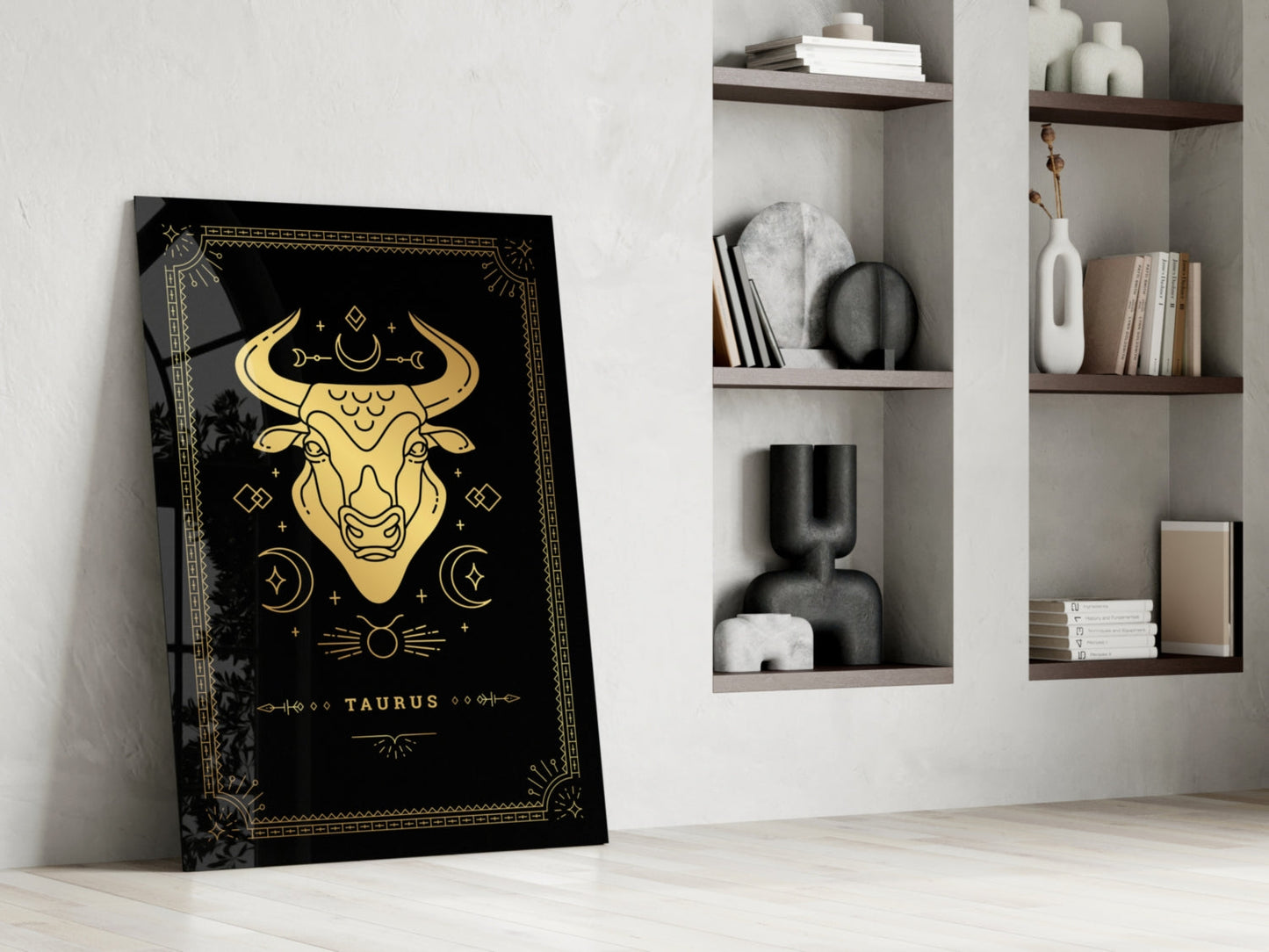 Black Taurus glass wall art with gold zodiac bull illustration leaning against white wall beside shelving unit.