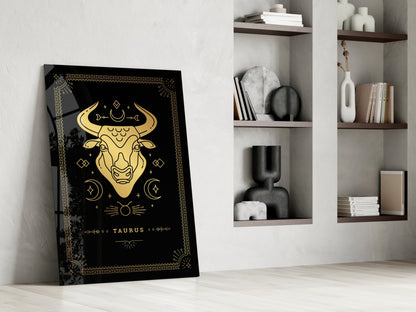 Black Taurus glass wall art with gold zodiac bull illustration leaning against white wall beside shelving unit.