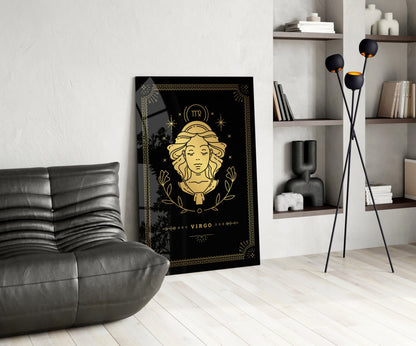 Black glass Virgo zodiac wall art with gold illustration displayed in modern living room beside grey chair and wooden shelving.