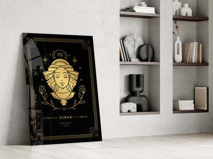Black glass Virgo zodiac wall art with gold illustration leaning against white wall beside shelving unit.