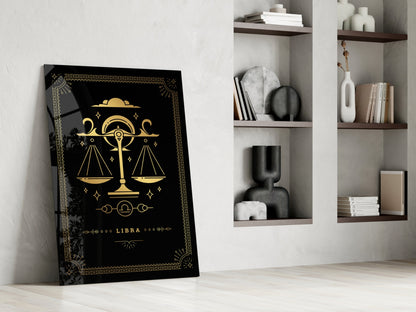 Black glass Libra zodiac wall art with gold scales and celestial symbols leaning against white wall beside shelving unit.